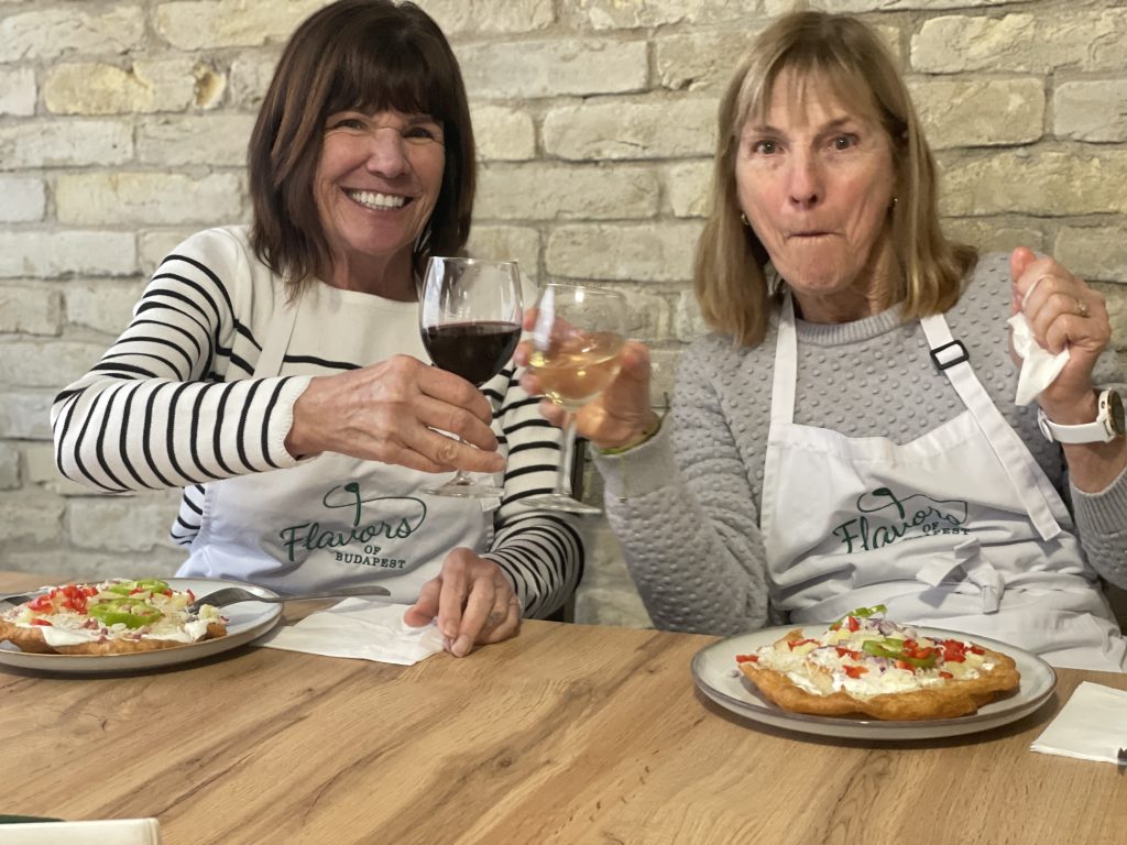 Two guests toasting with wine while enjoying classic and potato langos with toppings during a Hungarian cooking class.